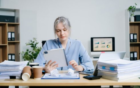 Contabilă analizând documente fiscale și calculând impozitul pe profit pe tabletă înconjurată de rapoarte financiare