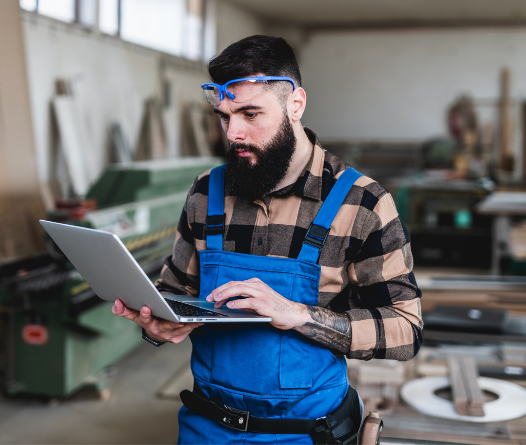 Muncitor cu salopetă albastră și ochelari de protecție folosind laptopul într-un atelier, ilustrând procesul simplu de obținere a creditului în 3 pași: creare cont, evaluare dosar și primirea fondurilor